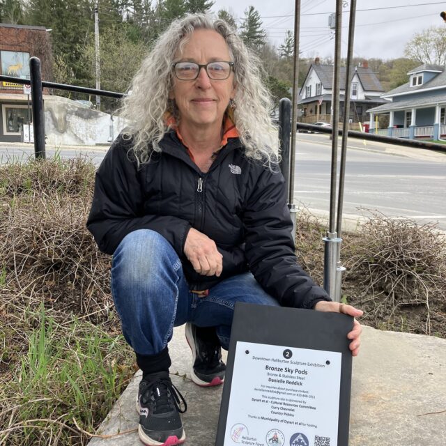 Danielle pictured with sponsor sign in Haliburton Sculpture Garden with her piece, Pods in the Sky